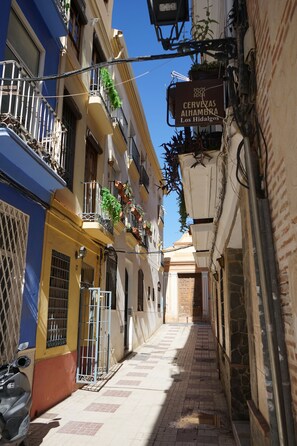 Una calle estrecha flanqueada por edificios, un letrero que dice 'Cervezas Alhambra Los Hidalgos' y una moto estacionada a la izquierda.