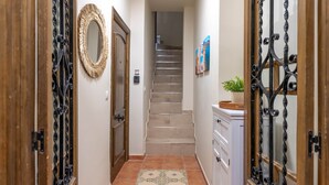Un pasillo con una escalera que conduce a un piso superior, una puerta con un espejo decorativo y un armario blanco con una cesta encima.