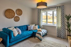 A living room with a blue sofa, patterned cushions, a woven wall art piece, a large window with curtains, and a potted plant. - Tir Ha Mor (Upton)