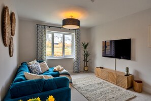 A living room with a sofa, a TV, a potted plant, and a window with curtains. - Tir Ha Mor (Upton)