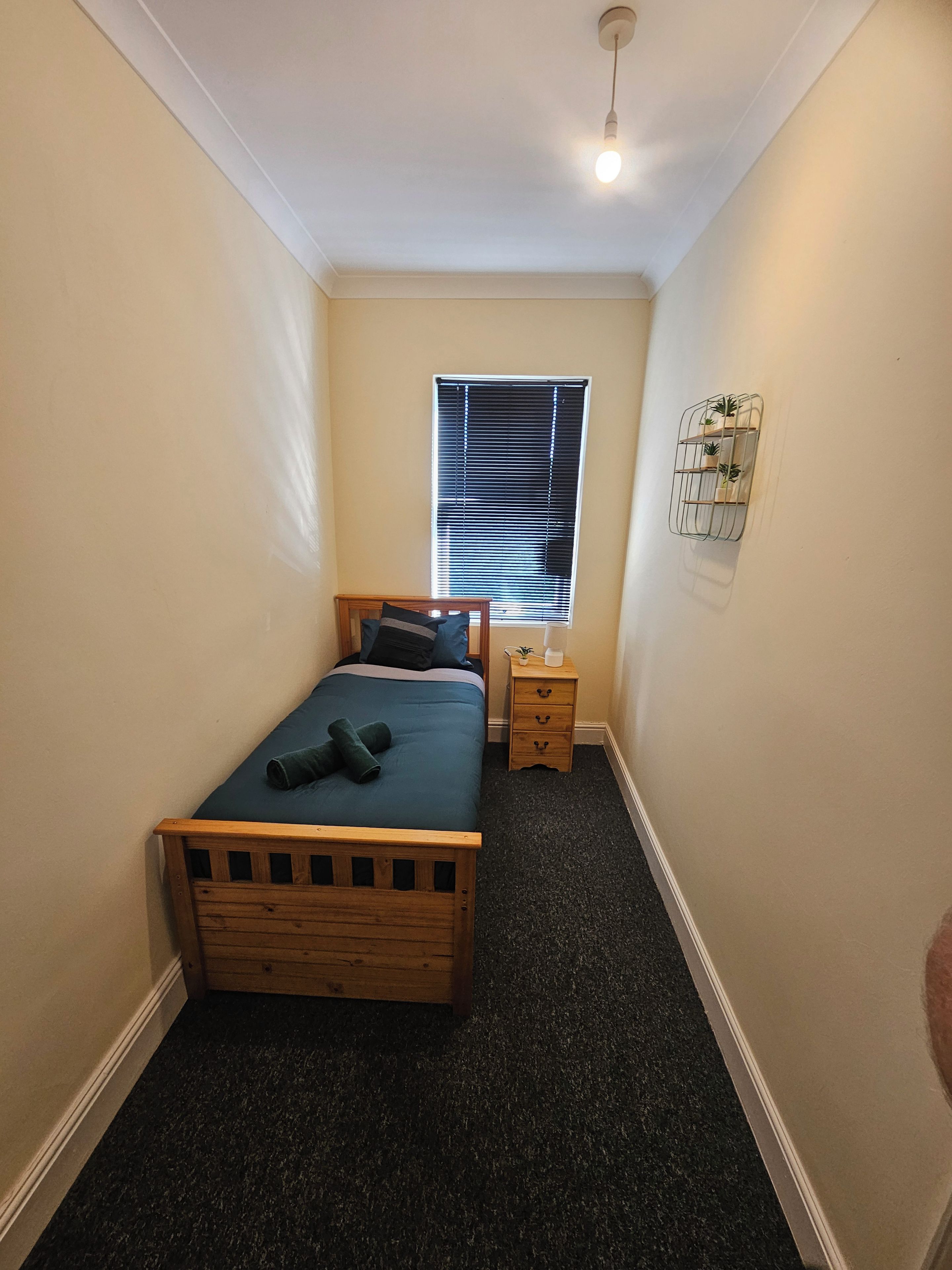 Ein Schlafzimmer mit einem Bett, Nachttischen, einem Fenster mit Jalousien und einem Wandregal mit Pflanzen.