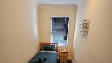 Ein Schlafzimmer mit einem Bett, Nachttischen, einem Fenster mit Jalousien und einem Wandregal mit Pflanzen.