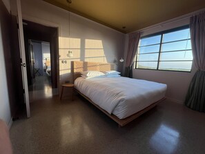 A bedroom with a bed, a bedside table, a window with curtains, and a door leading to another room.