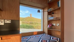 A cozy room with a built-in bench, wooden shelves, and a window offering a view of a hill and a tower.