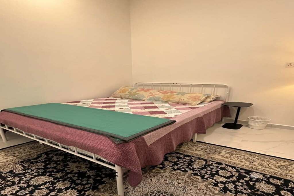 A single-bed room with a metal frame, a patterned bedspread, a green throw blanket, a small bedside table, and a patterned rug.