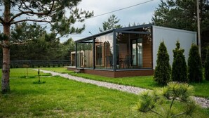 Una cabana moderna amb tancament de vidre, terrassa de fusta i un jardí envoltant.