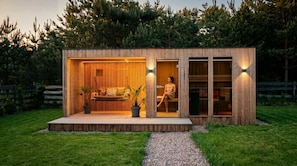 A modern wooden cabin with a glass door entrance, a small outdoor seating area, and a gravel path leading to the door.