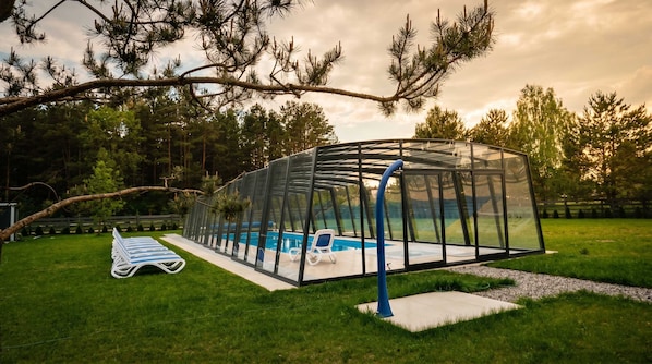 A covered swimming pool with a glass enclosure, surrounded by a grassy yard and trees.