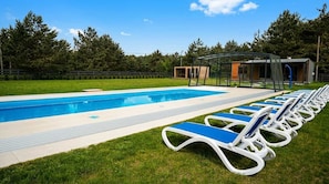 A swimming pool with lounge chairs and a covered area, surrounded by grass and trees.