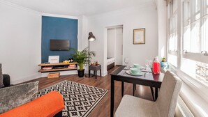 Een moderne woonkamer met een flatscreen televisie, een eettafel gedekt voor twee personen, een potplant en een groot raam dat natuurlijk licht binnenlaat.