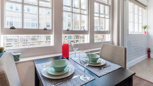 Een eetgedeelte met een tafel voor twee personen, grote ramen met uitzicht op gebouwen, een kamerplant op een plank en een brandblusser aan de muur.