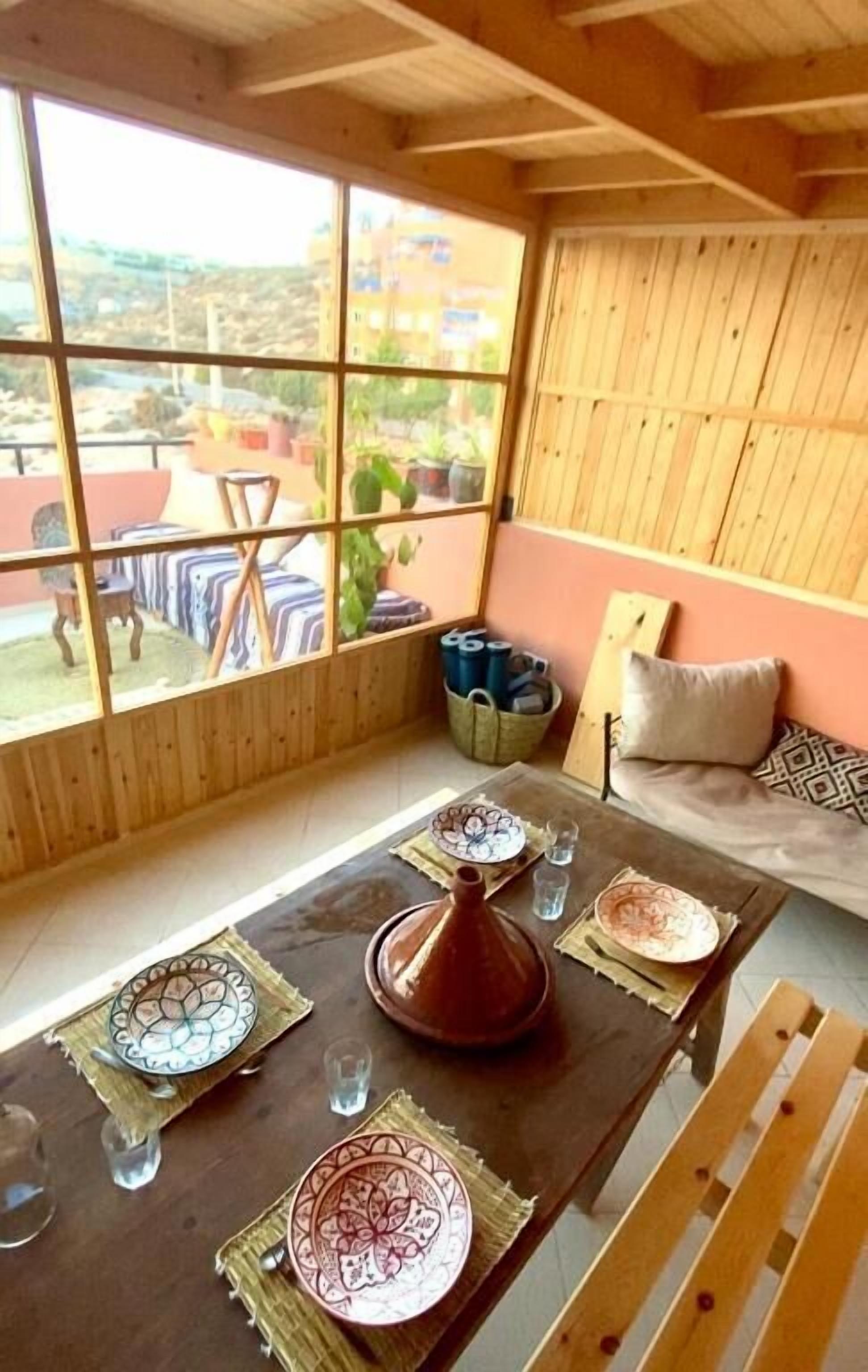 Un interior de madera con una mesa de comedor preparada para dos, un sofá y vistas a través de amplios ventanales.