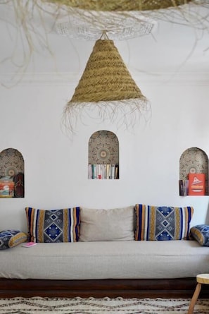 A room with a sofa, patterned cushions, a woven hanging light fixture, and decorative wall art. - Moana Surf Maroc (Aourir)