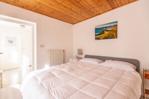 A bedroom with a large bed, a wooden ceiling, a radiator, a bedside table with a lamp, and a painting on the wall.