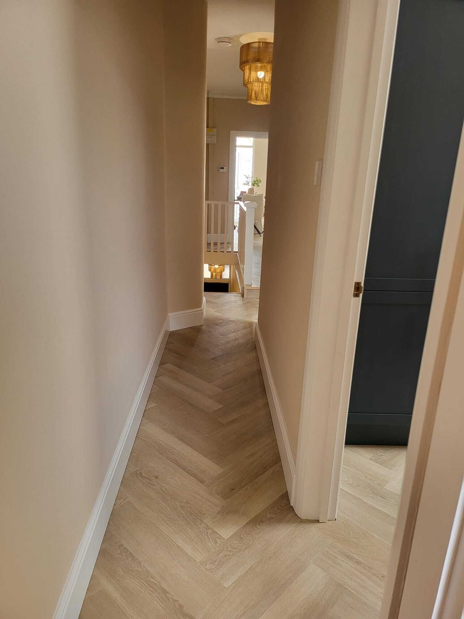 Un pasillo con un patrón de suelo tipo espiga que conduce a una escalera y una habitación con una puerta oscura.