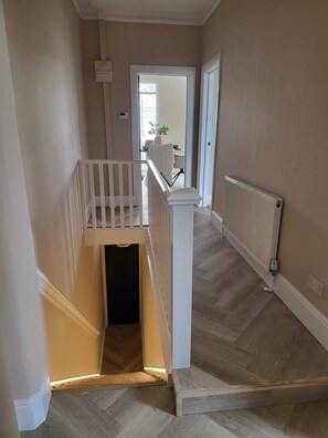 A staircase with a white railing leading to a landing, a radiator on the wall, and a door at the end of the hallway.