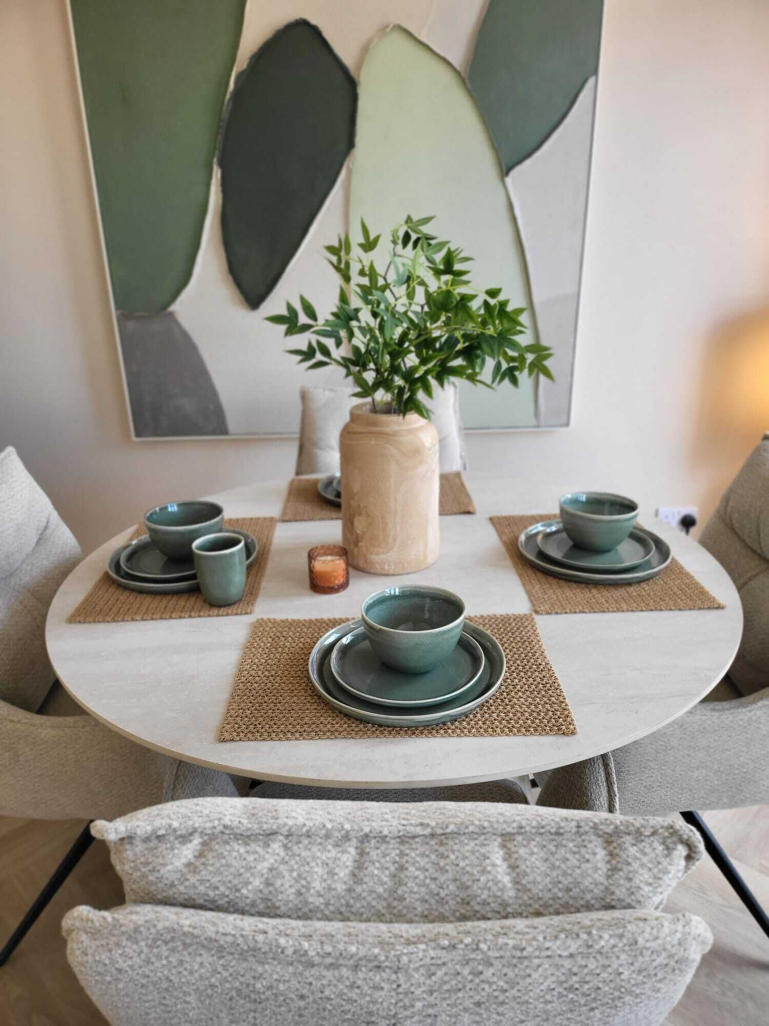 Une table de salle à manger ronde, ornée d’un vase rempli de verdure, de deux jeux de bols et d’assiettes assortis, et d’un set de table texturé.
