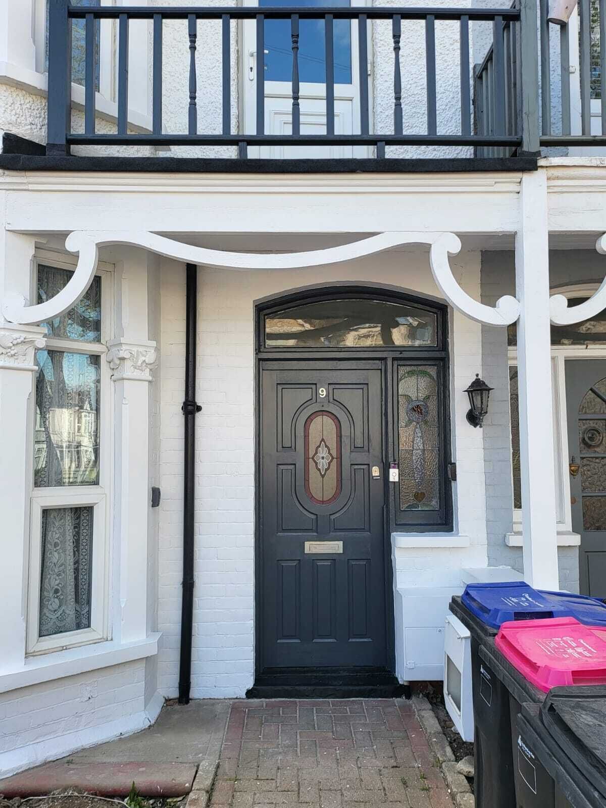 A two-story building with a black front door, white exterior walls, and a balcony with black railings.