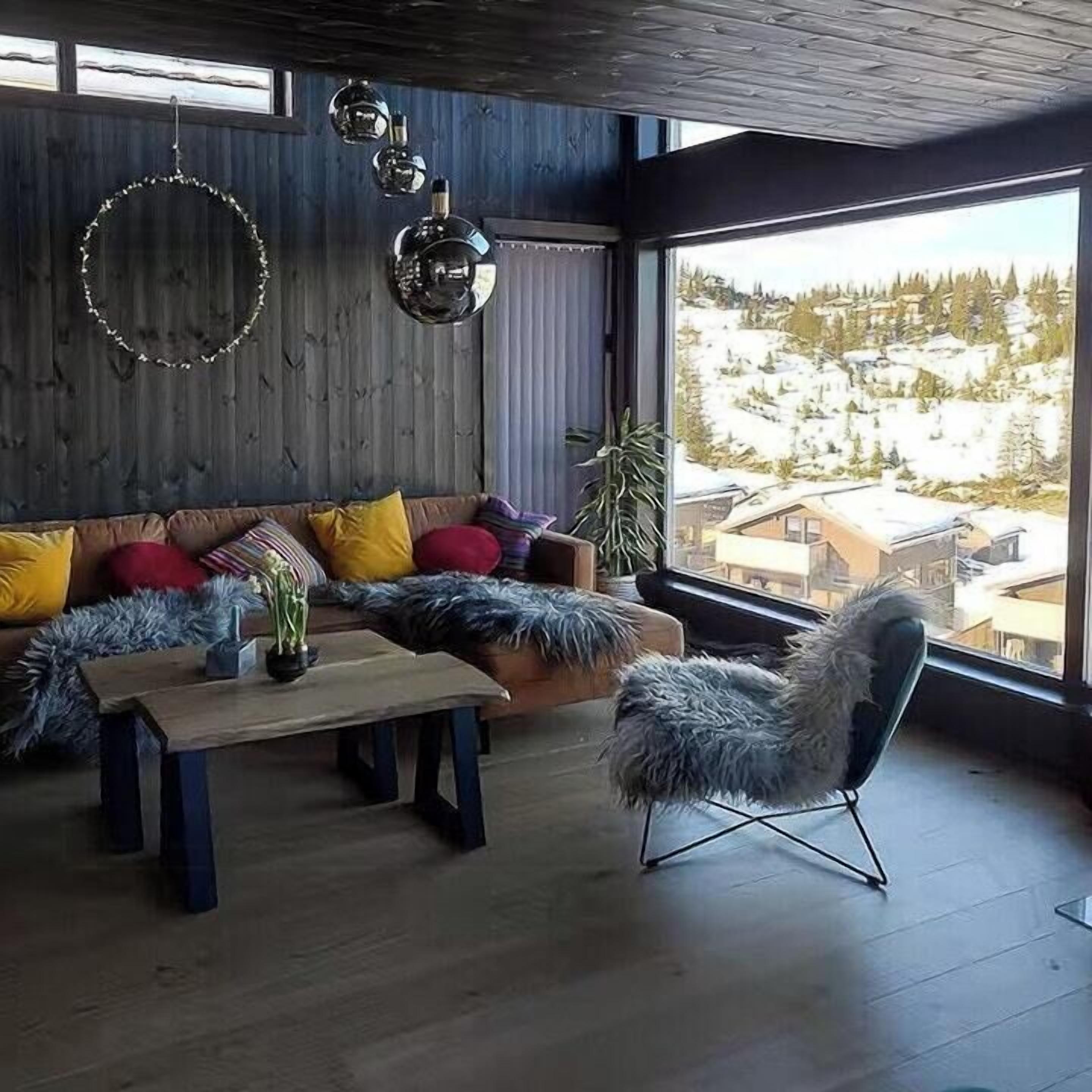 A cozy room with a wooden floor, a sofa with colorful cushions, a round mirror, and a view of a snowy landscape.