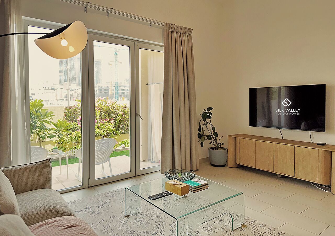 Una sala de estar moderna con una mesa de centro de vidrio, un sofá, un televisor y un ventanal corredizo que da a un espacio exterior.