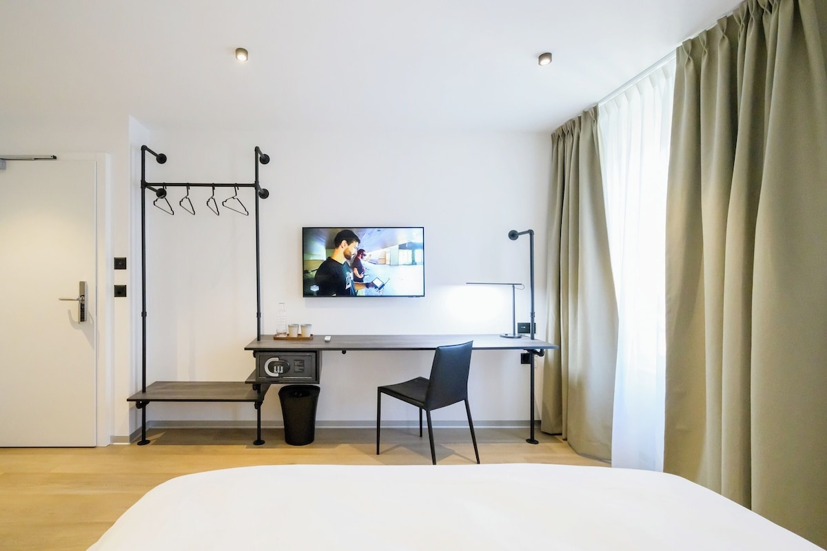 Ein Hotelzimmer mit Bett, Schreibtisch mit Stuhl, Wandfernseher und einem Fenster mit Vorhängen.