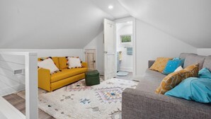 A loft-style bedroom with a high ceiling, a yellow sofa, a grey sofa, and a patterned rug.