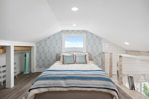 Ein Schlafzimmer mit schrägverdeckter Decke, einem Bett mit gestreifter Decke, einem Fenster mit Ausblick und einer eingebauten Garderobe.