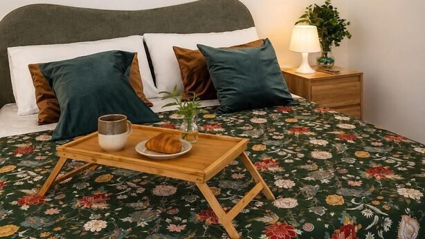 A hotel room with a bed, pillows, a bedside table with a lamp, a tray with a croissant and a cup, and two wall art pieces.