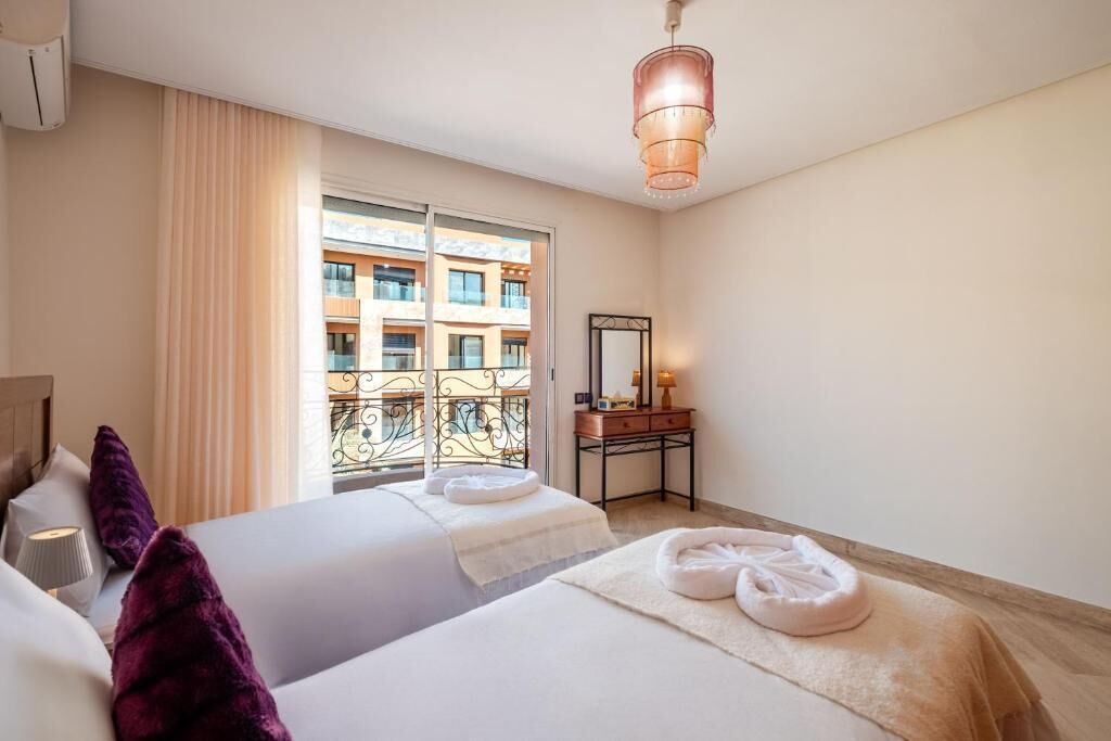 Habitación de hotel con dos camas, una ventana con vistas a un edificio, una mesita de noche con lámpara, y un candelabro colgante.