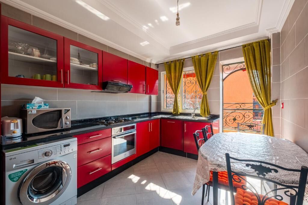 A compact kitchen with red cabinets, a microwave, a washing machine, and a dining area with a table and chairs.