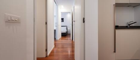 A hallway with wooden flooring leading to a bedroom with a bed and nightstand.
