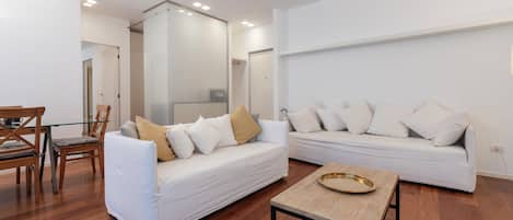 Una sala moderna con una mesa de comedor de vidrio, dos sofás blancos con cojines y una mesa de centro de madera.