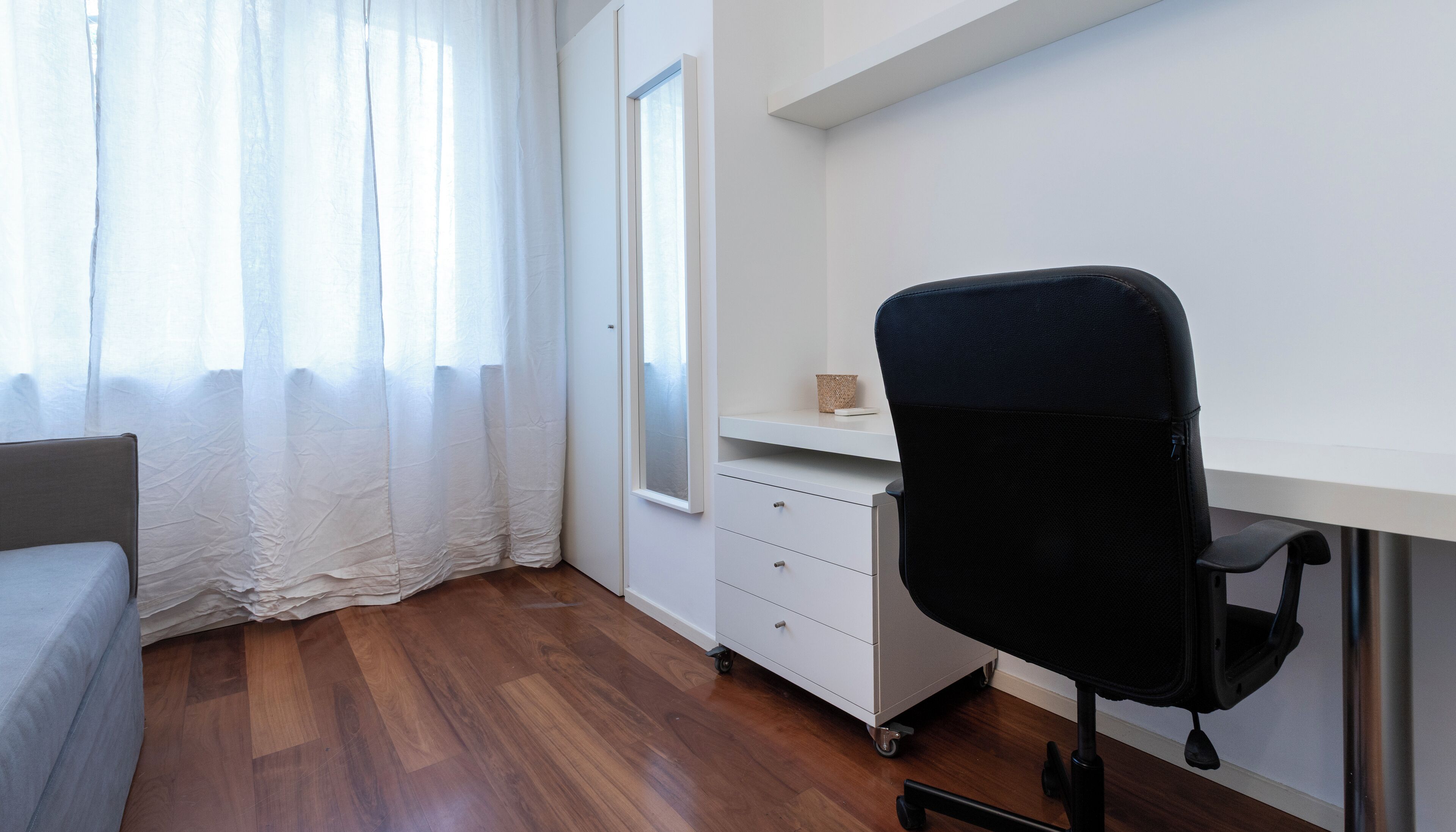 Ein Zimmer mit Holzboden, einem weißen Schreibtisch mit Stuhl, einem weißen Schrank und einem Fenster mit durchsichtigen Vorhängen.