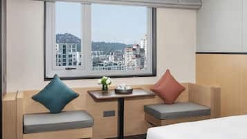 A hotel room with a bed, a small table with a vase, two cushioned seats, and a window with a city view.