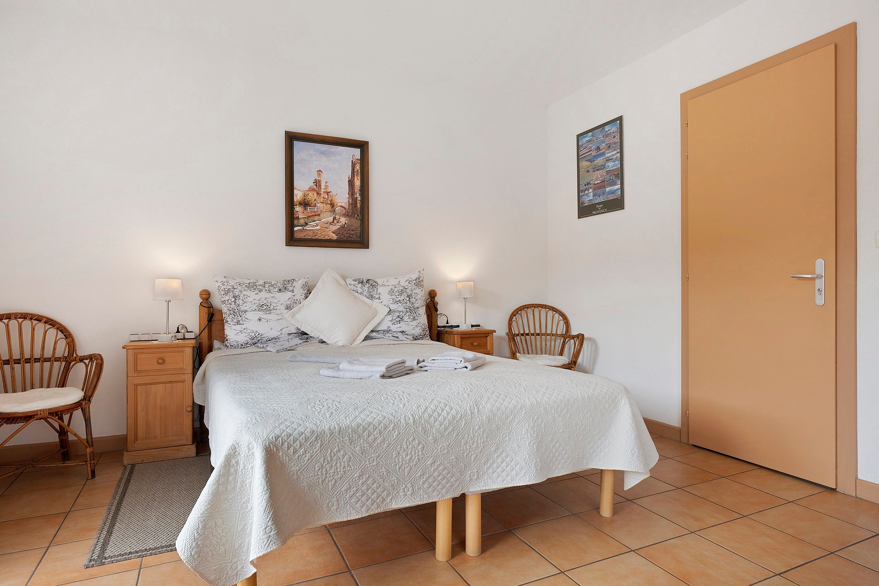 A neatly arranged bedroom with a bed, two bedside tables, two chairs, a door, and two wall-mounted pictures.