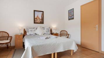 A neatly arranged bedroom with a bed, two bedside tables, two chairs, a door, and two wall-mounted pictures.