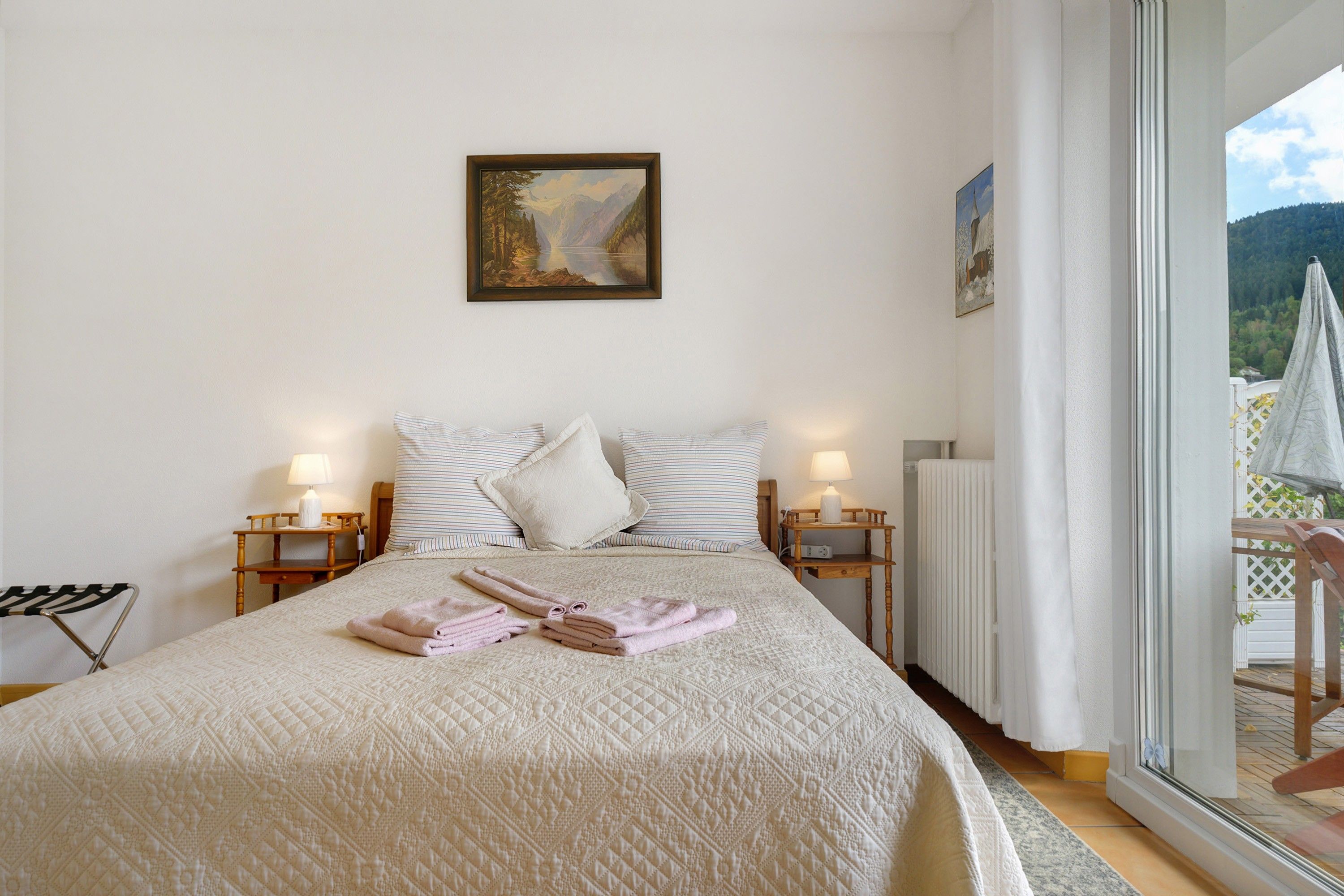 A bedroom with a bed, pillows, a painting, bedside lamps, and a view of a balcony with umbrellas.