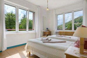 Une chambre d’hôtel avec deux lits, un bureau, une chaise, une lampe et de grandes fenêtres donnant sur un paysage verdoyant et vallonné.