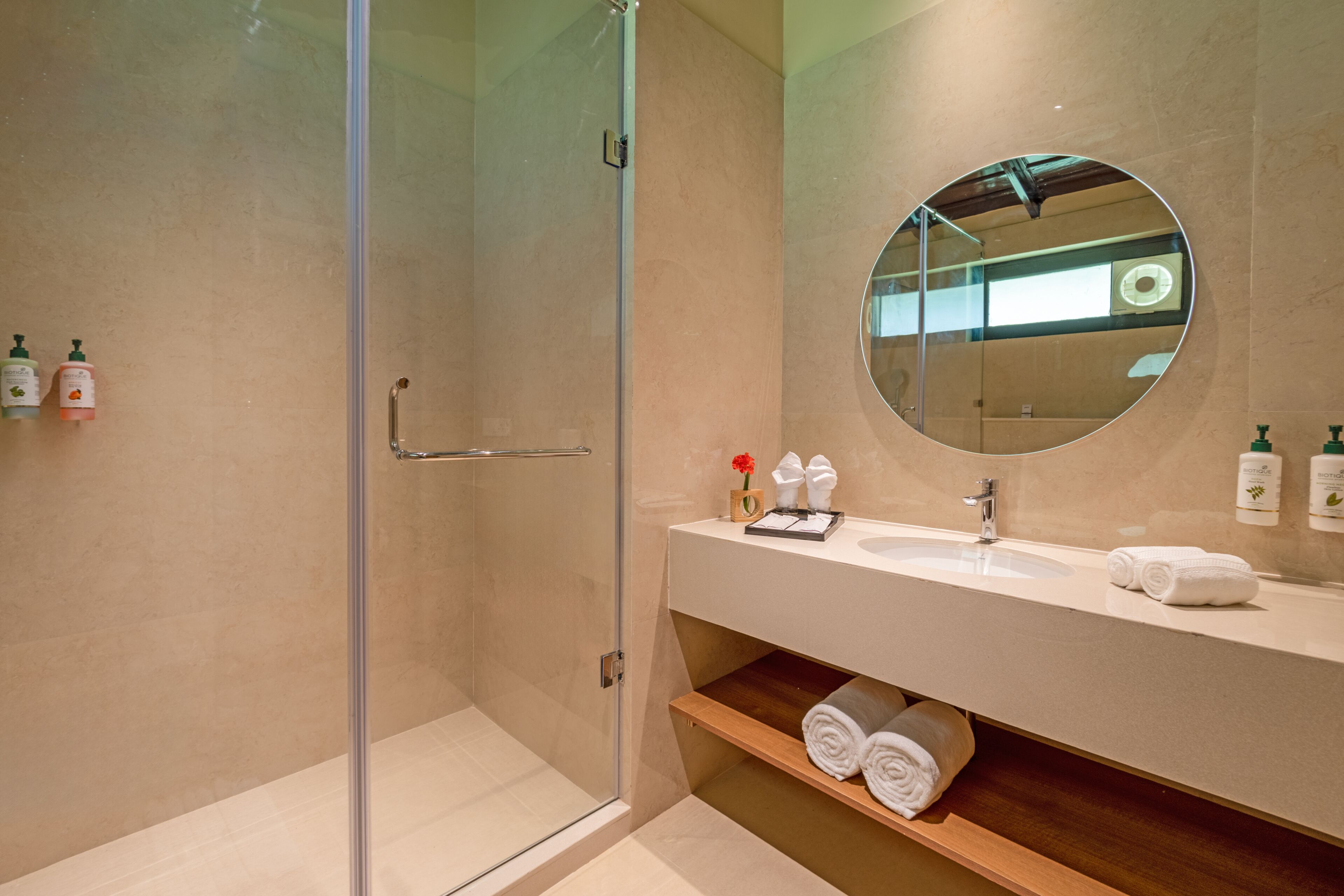 A modern bathroom with a glass-enclosed shower, a round mirror, and a white sink with toiletries.