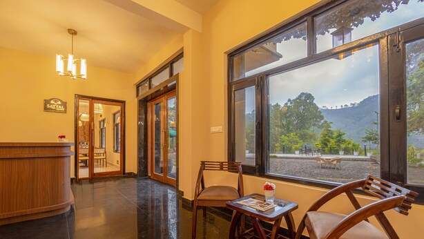 Interior de un restaurante con mostrador de madera, zona de comedor con sillas y un amplio ventanal con vistas a un jardín y colinas.