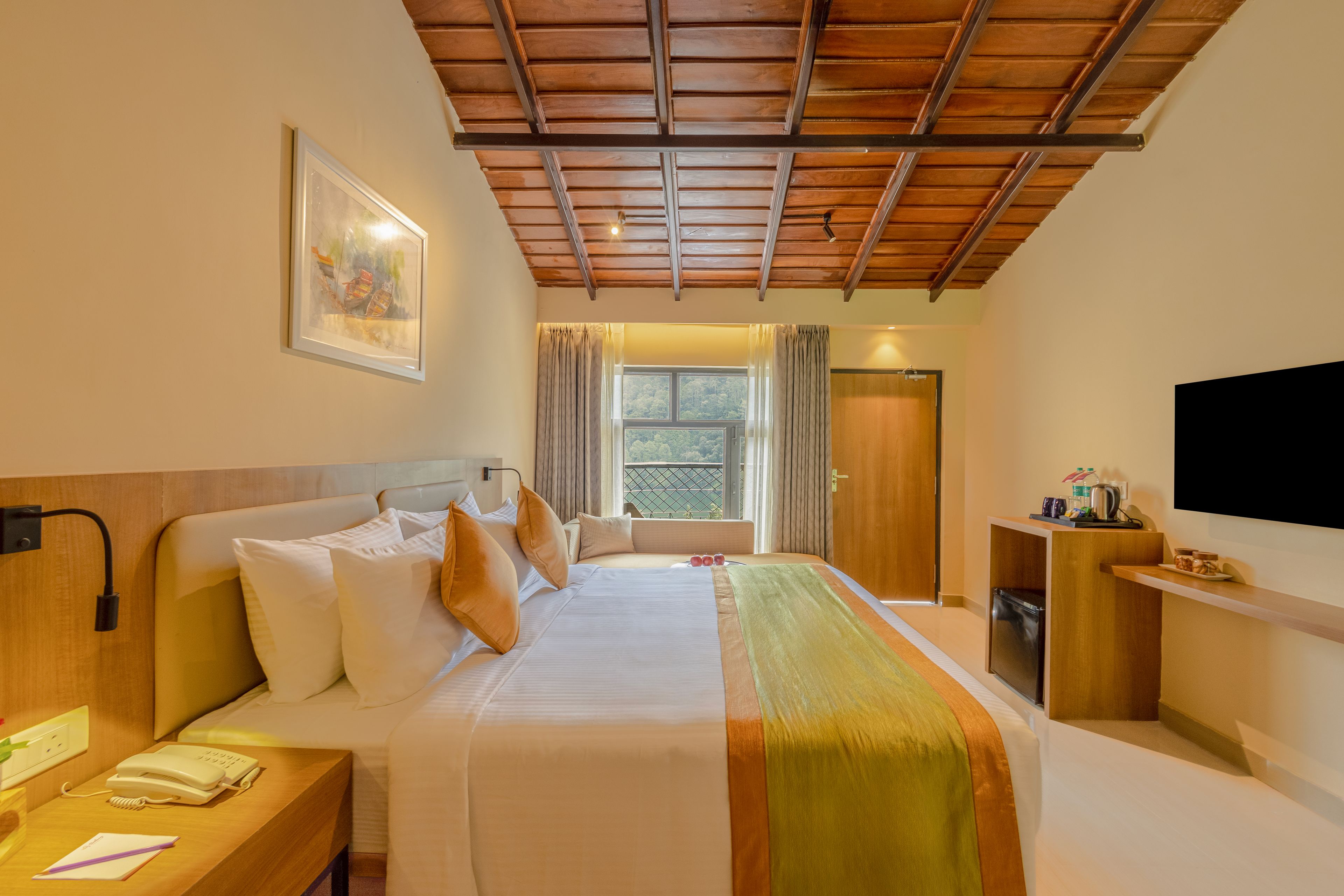 Een hotelkamer met een bed, nachtkastje, schilderij, raam met gordijnen, televisie en een klein kitchenette.