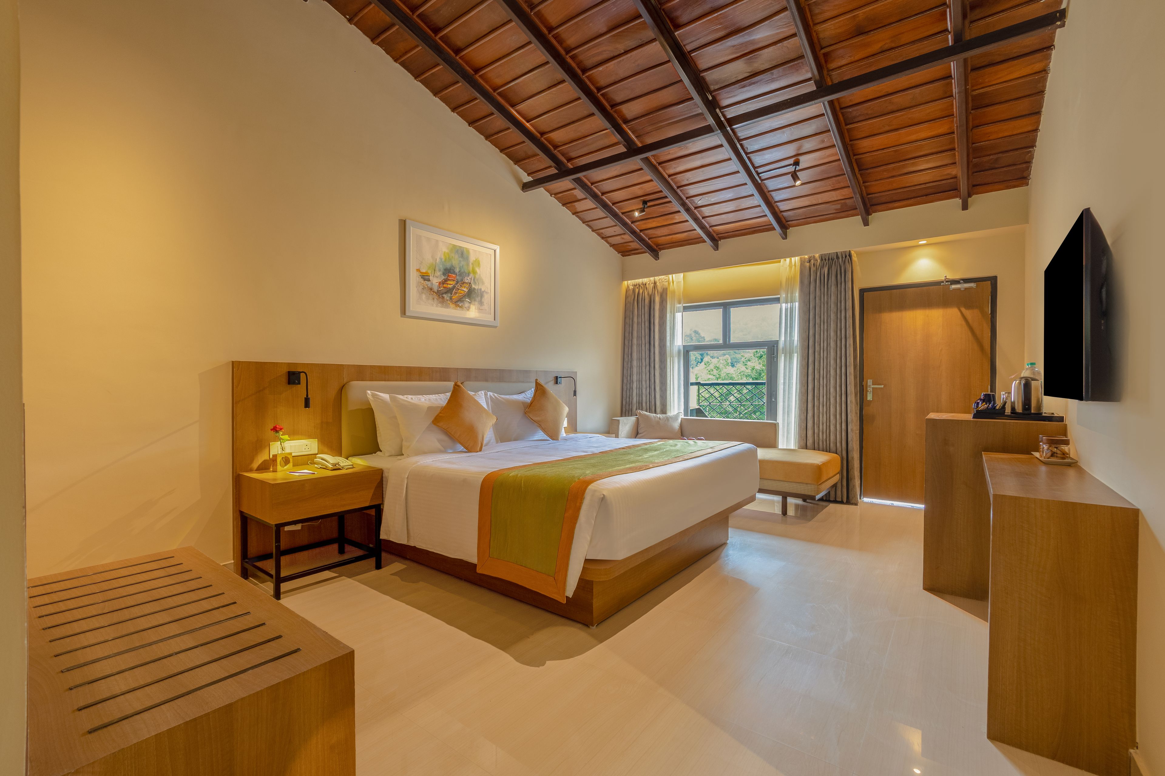 A hotel room with a wooden ceiling, a bed with a green and orange bedspread, a bedside table with a lamp, a painting on the wall, and a TV on the wall.