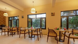 Un comedor con varias mesas y sillas, ventanas amplias con vista a árboles e iluminación colgante.