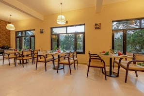 Un comedor con varias mesas y sillas, ventanas amplias con vista a árboles e iluminación colgante.