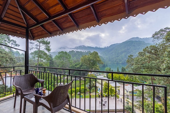 A balcony with wicker furniture and a table, overlooking a mountainous landscape with a view of a lake and greenery.