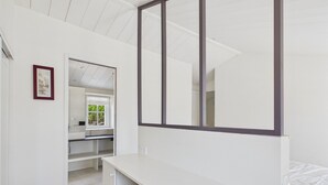 Ein Schlafzimmer mit einem weißen Bett, einem Schreibtisch und einem Stuhl. Ein Fenster bietet Blick auf grüne Vegetation.