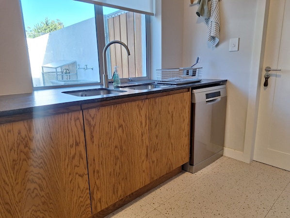 A kitchen with wooden cabinets, a stainless steel dishwasher, and a window with a view of a building outside. - 6 on Heide (Franschhoek)
