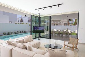 A modern living room with a pool, a flat-screen TV, and a dining area with outdoor seating. - Ocean Whispers 2, New Villa with private pool, within 100 meters from the beach (Protaras)