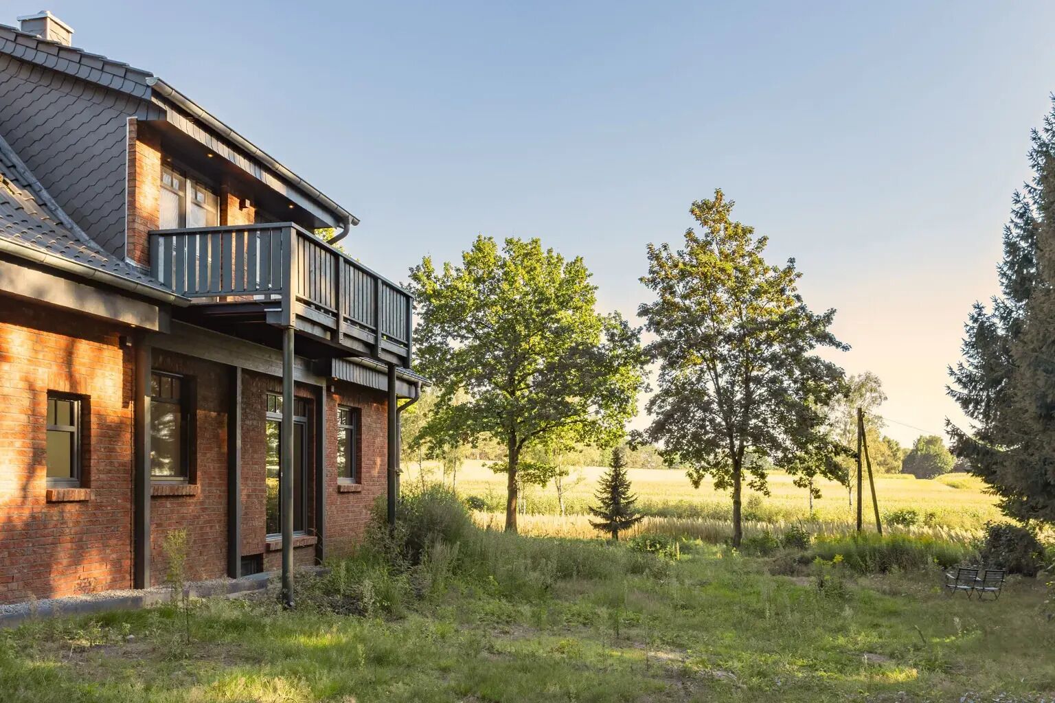 A modern house with a brick exterior, a balcony, and a view of a grassy field with trees.
