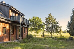Uma casa moderna com fachada de tijolos, uma varanda e vista para um campo gramado com árvores.